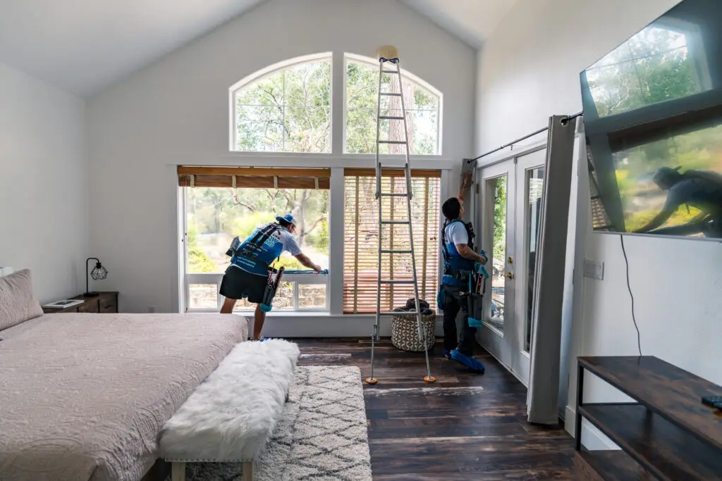 Two professional window cleaners in a bright, modern bedroom, one cleaning a large window while the other prepares to clean a door, showcasing meticulous work and attention to detail.